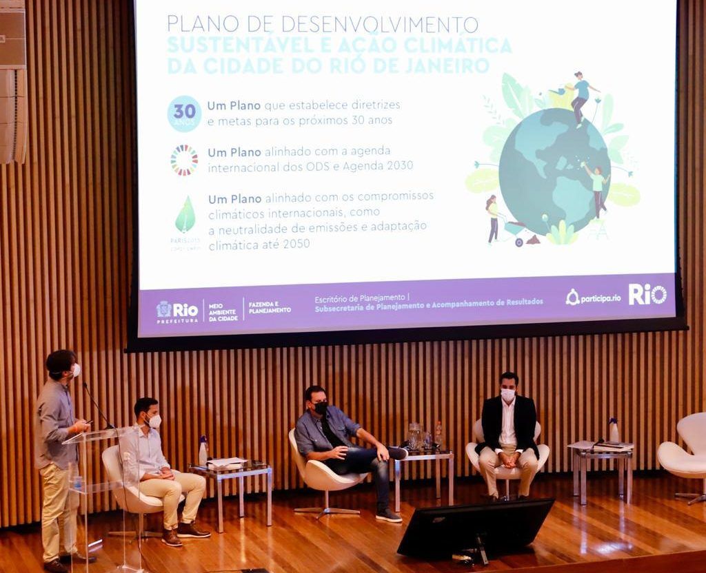 Quatro responsáveis pelo Plano de Desenvolvimento Sustentável e Ação Climática discutem os objetivos da ação, que foi lançada por meio da Prefeitura do Rio de Janeiro, a partir da apresentação em um telão para todos os presentes no evento.